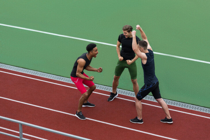 Picture of happy athlete team standing on running track outdoors make winner gesture